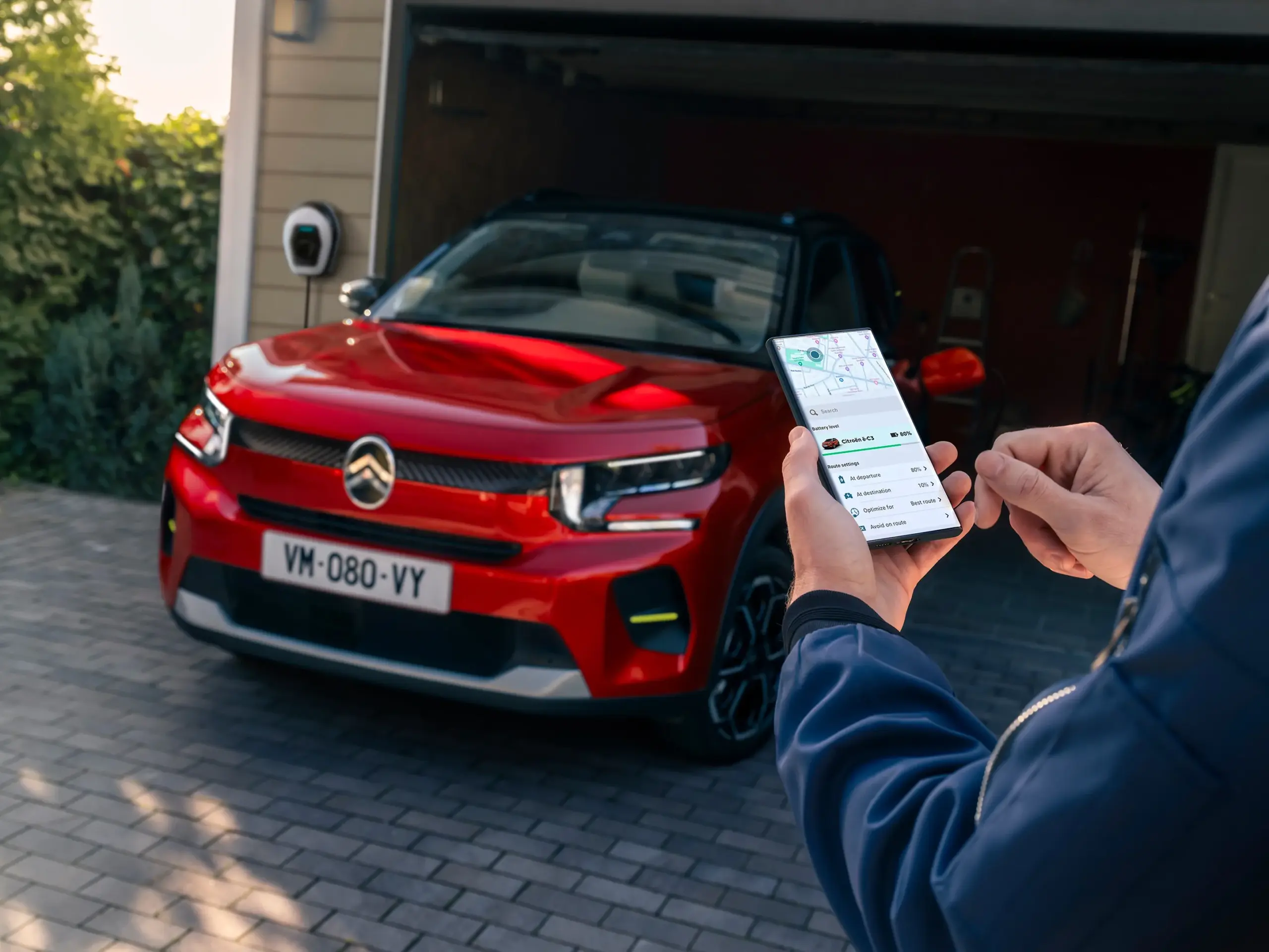 Roter Citroën ë-C3 an einer Ladestation, gesteuert über eine Smartphone-App vor einer Garage.