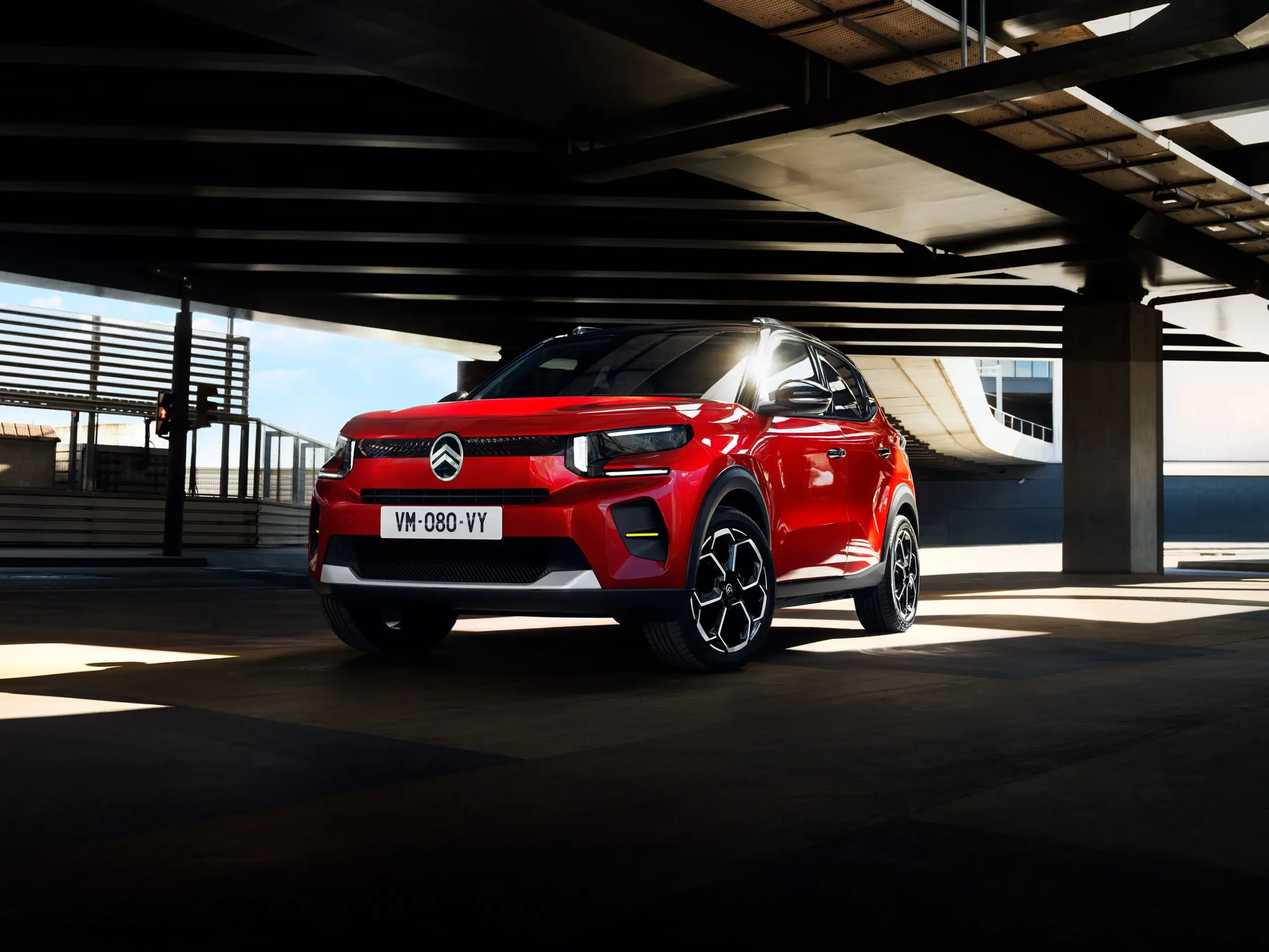 Citroën ë-C3 in Rot, frontal fotografiert, unter einem Parkhaus, modernes urbanes Umfeld.