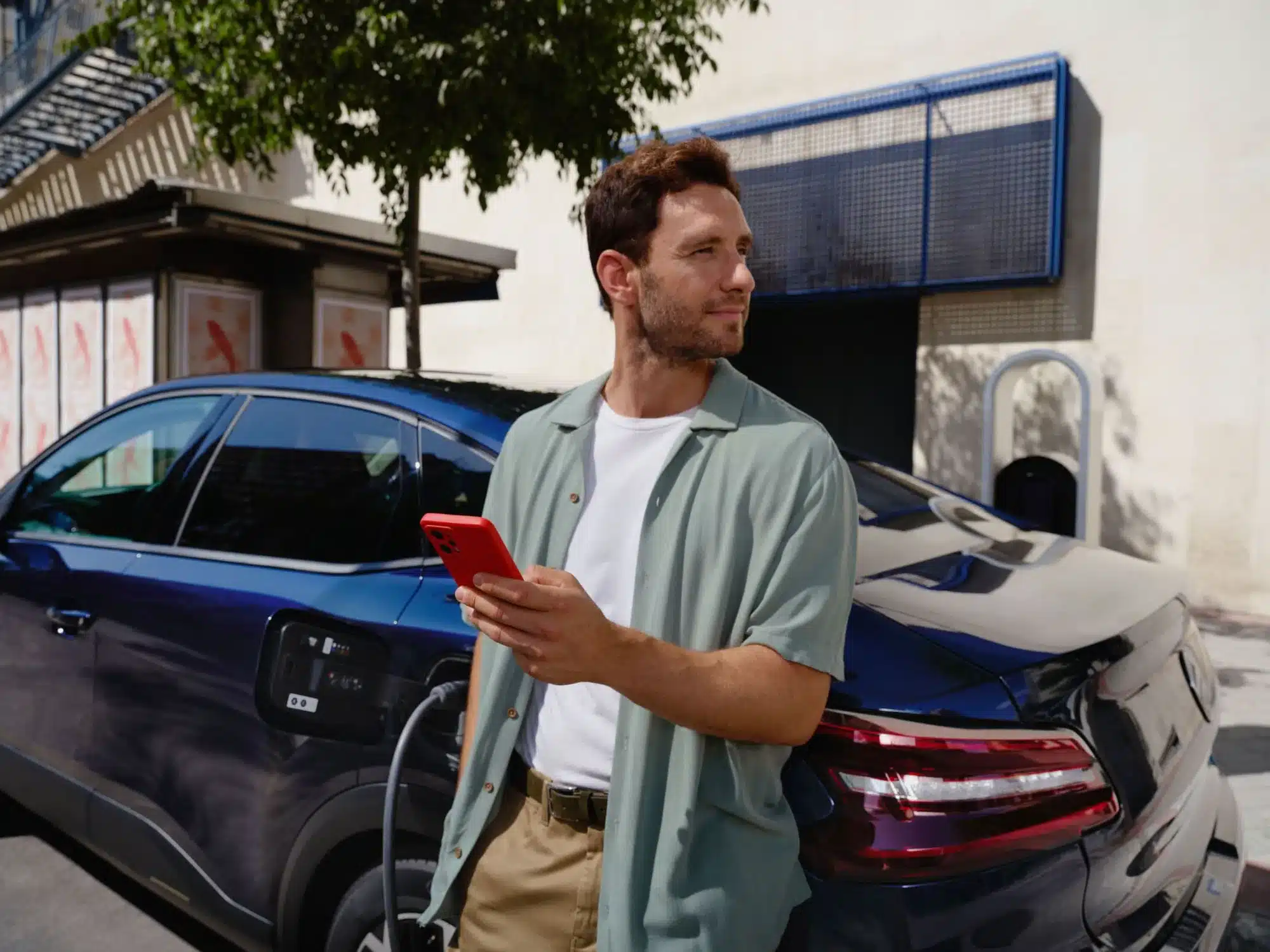 Citroën New C4 X wird an einer öffentlichen Ladesäule geladen; ein Mann steht daneben mit Smartphone.