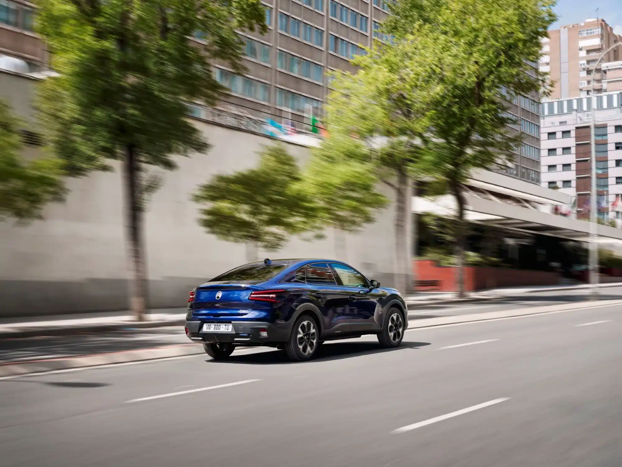 Blauer Citroën New C4 X fährt auf einer städtischen Straße entlang, Fokus auf der eleganten Seiten- und Heckpartie.