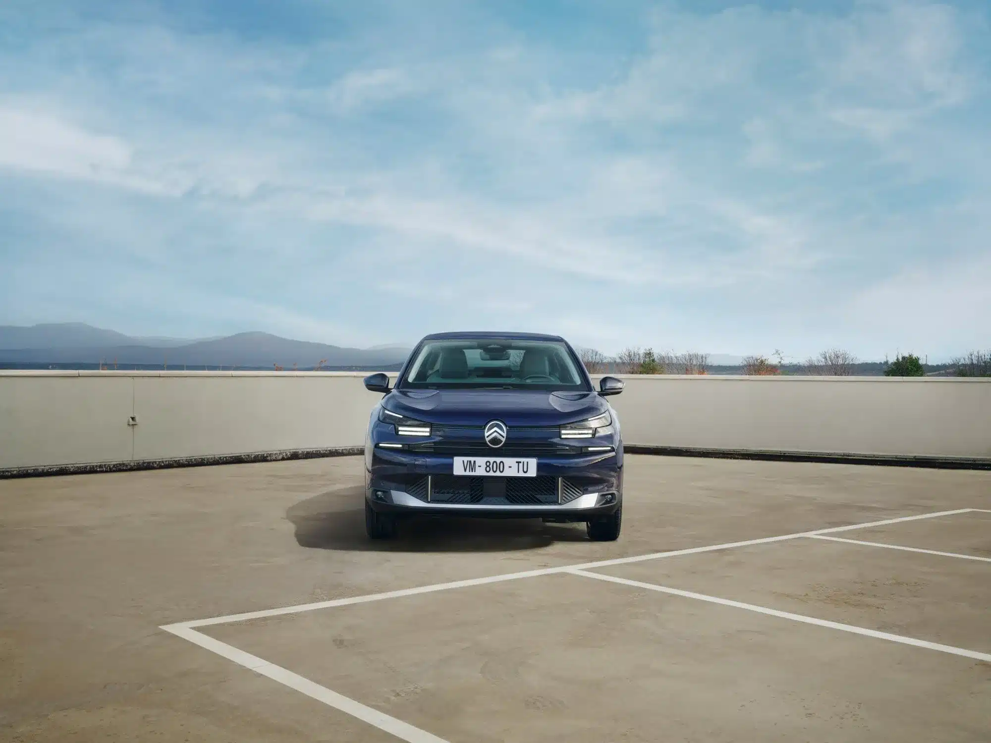 Citroën New C4 X von vorne, geparkt auf einem offenen Parkplatz mit Gebirgslandschaft im Hintergrund.