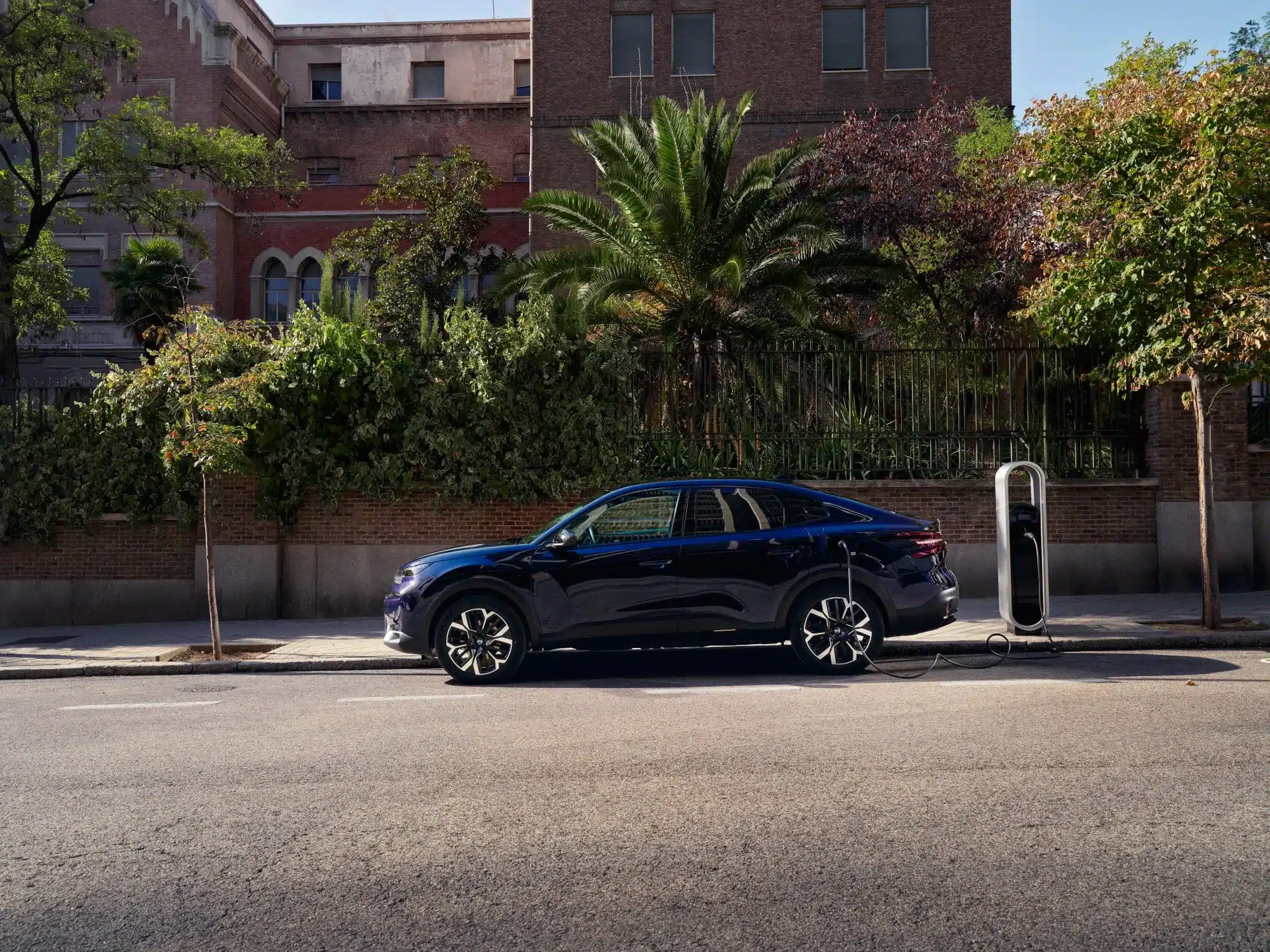 Seitenansicht des Citroën New C4 X an einer Ladestation vor einem Gebäude mit Grünanlage.