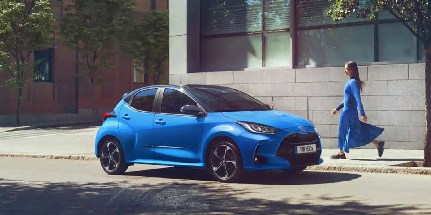 Blauer Toyota Yaris Hybrid geparkt am Straßenrand vor einem modernen Gebäude, eine Frau geht auf dem Gehweg vorbei.