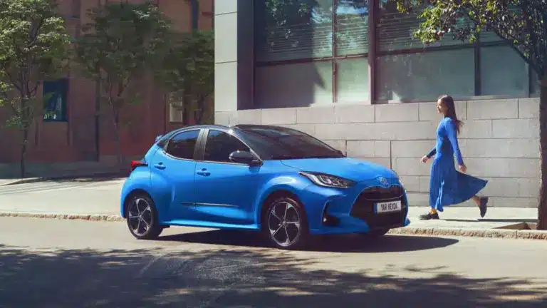 Blauer Toyota Yaris Hybrid geparkt am Straßenrand vor einem modernen Gebäude, eine Frau geht auf dem Gehweg vorbei.