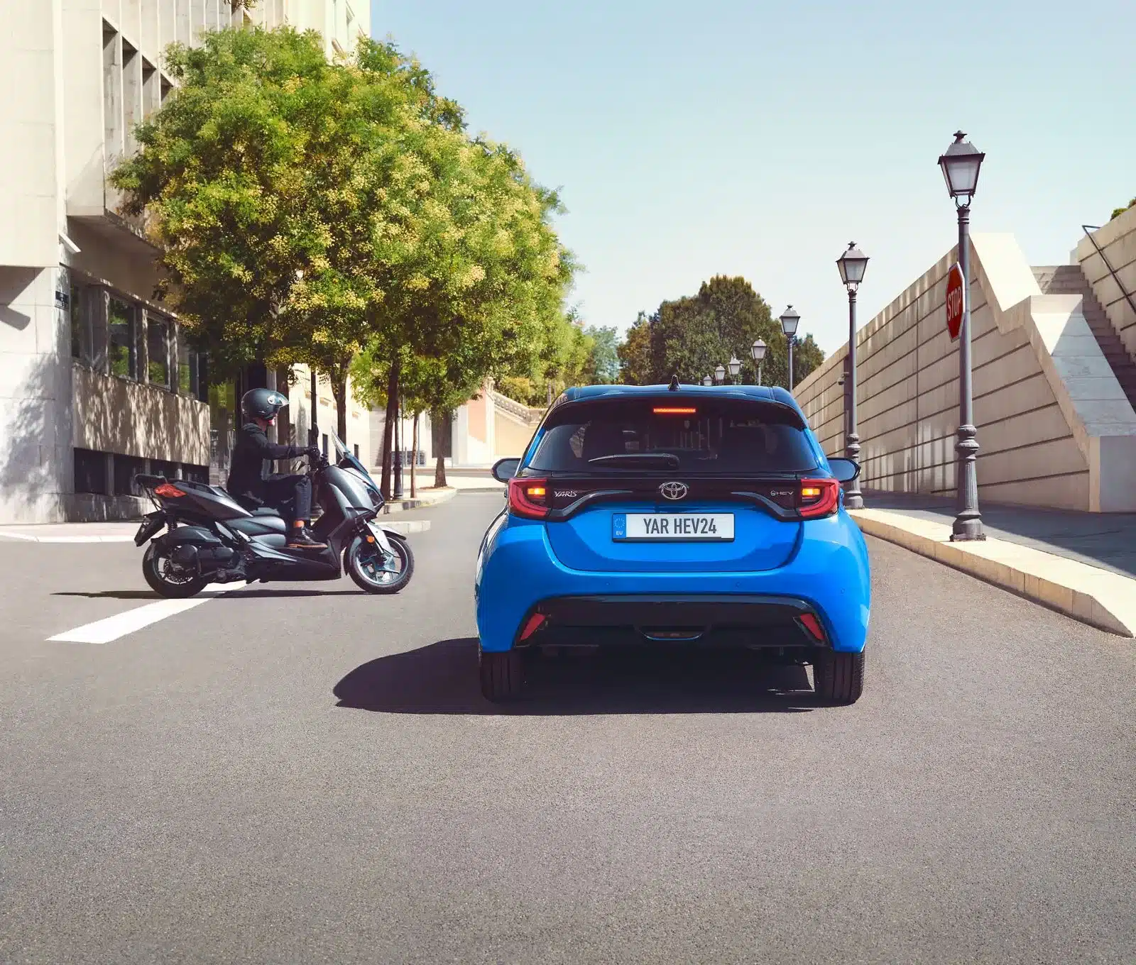 Blauer Toyota Yaris Hybrid von hinten, fährt in einer Stadtstraße, Motorradfahrer im Hintergrund.