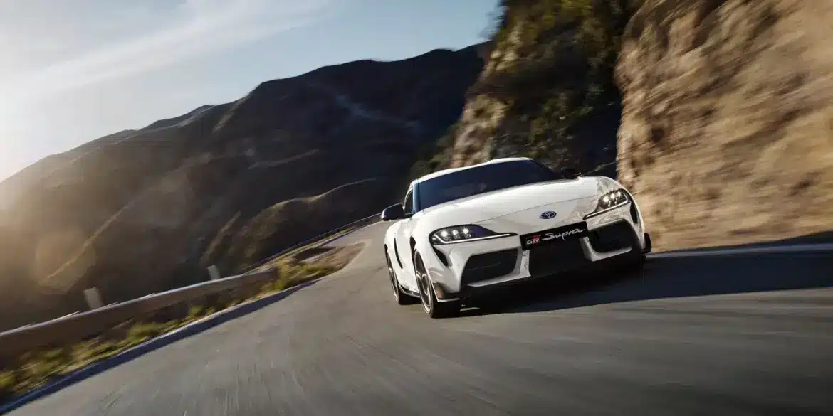 Weißer Toyota GR Supra 2025 fährt dynamisch auf einer kurvigen Straße in bergiger Landschaft.