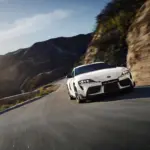 Weißer Toyota GR Supra 2025 fährt dynamisch auf einer kurvigen Straße in bergiger Landschaft.