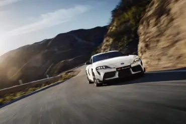 Weißer Toyota GR Supra 2025 fährt dynamisch auf einer kurvigen Straße in bergiger Landschaft.