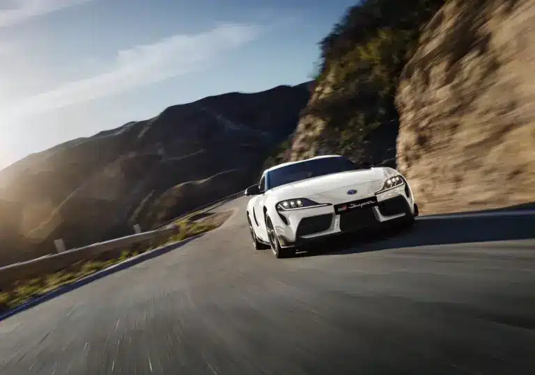Weißer Toyota GR Supra 2025 fährt dynamisch auf einer kurvigen Straße in bergiger Landschaft.