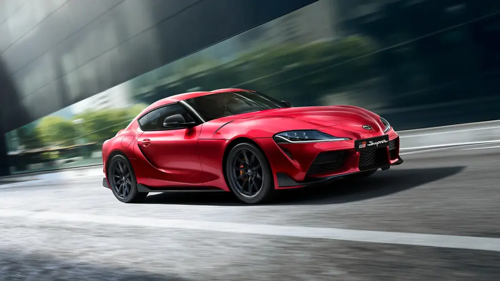 Roter Toyota GR Supra 2025 fährt sportlich und dynamisch auf der Straße vor moderner Architektur.