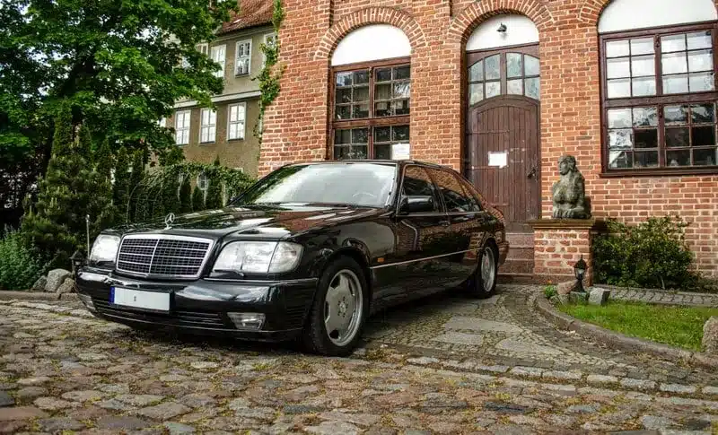 Dunkelgrüner Mercedes-Benz 600 SEL (W140) von 1991 parkt auf einem historischen Kopfsteinpflaster vor einer Villa.