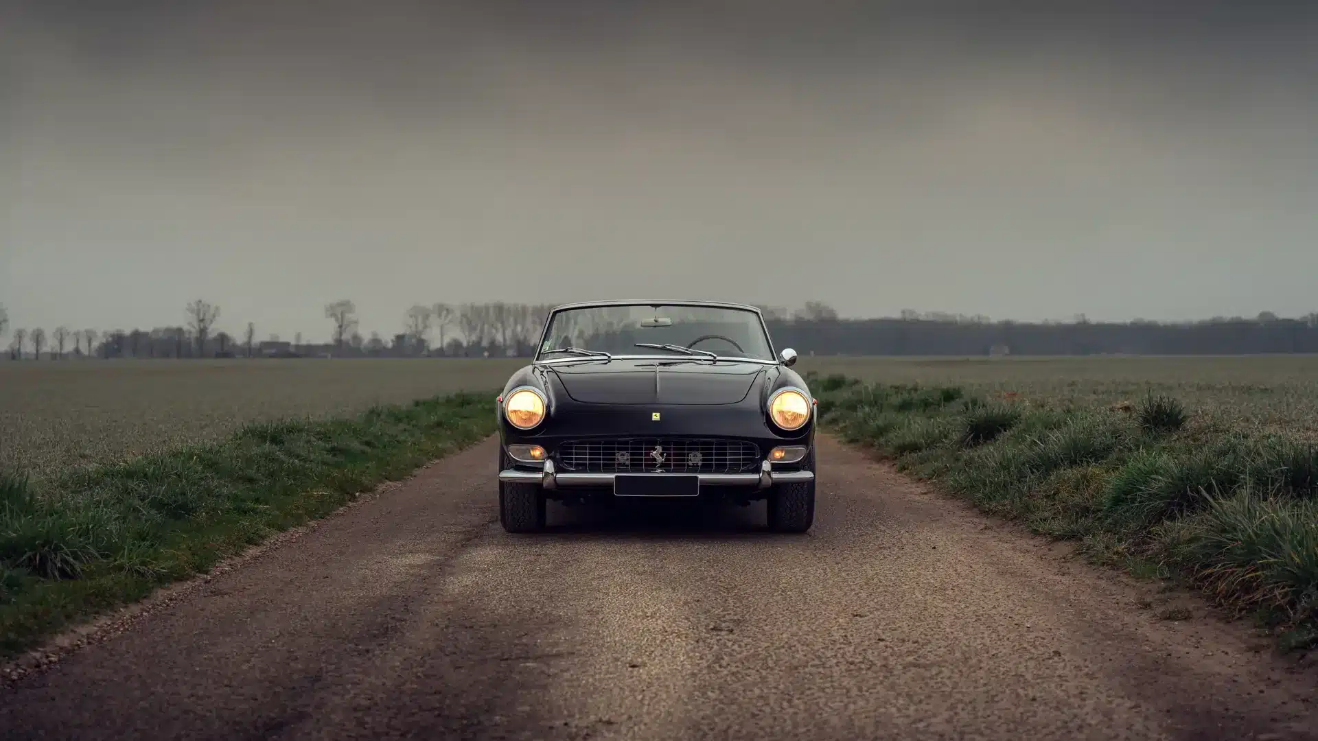 Ferrari 275 GTS von vorne, Schwarz, Scheinwerfer eingeschaltet, ländliche Straße unter wolkenverhangenem Himmel.
