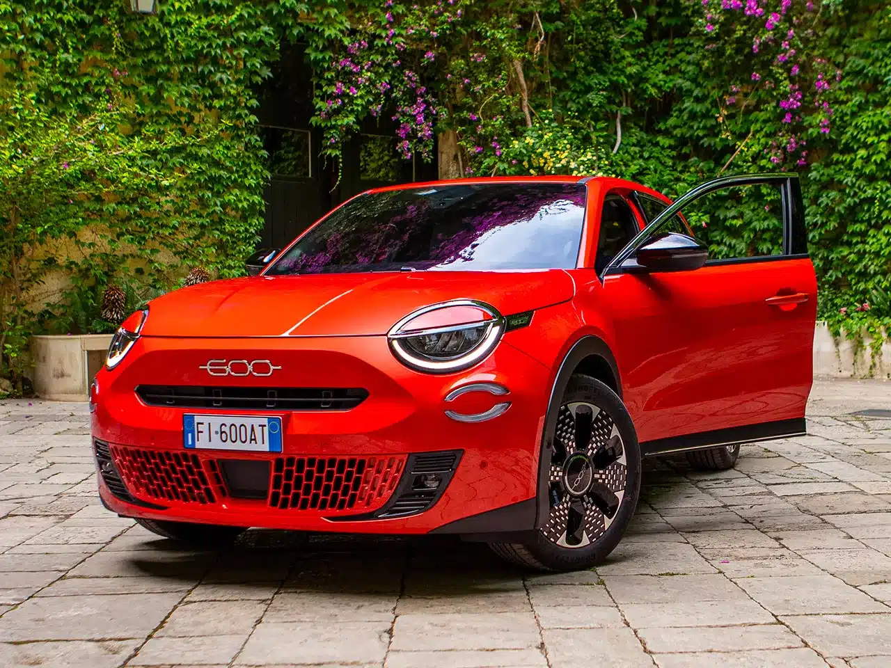 Roter Fiat 600 Elektro mit geöffnetem Fahrertür steht auf einem gepflasterten Hof vor einer Wand mit Efeu und Blumen.