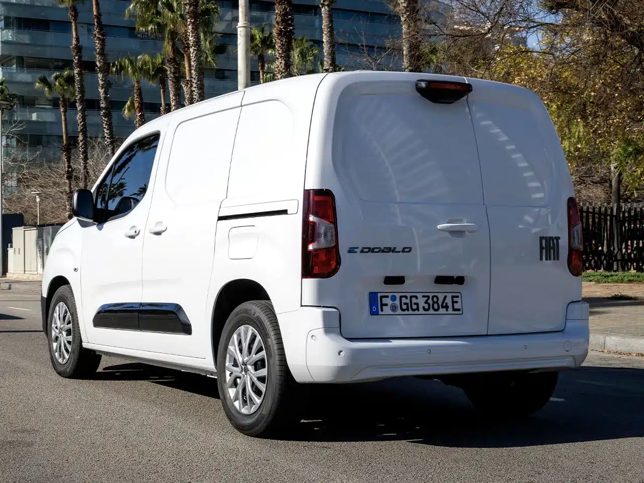 Heckansicht eines weißen Fiat Doblò 25th Anniversary Kastenwagens auf einer Straße, umgeben von Palmen und moderner Architektur.