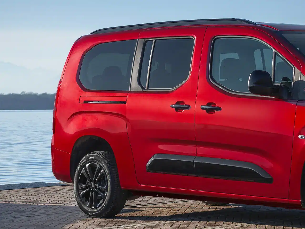 Roter Fiat Qubo L, seitliche Aufnahme mit großen Fenstern und Schiebetüren, vor Wasser stehend.