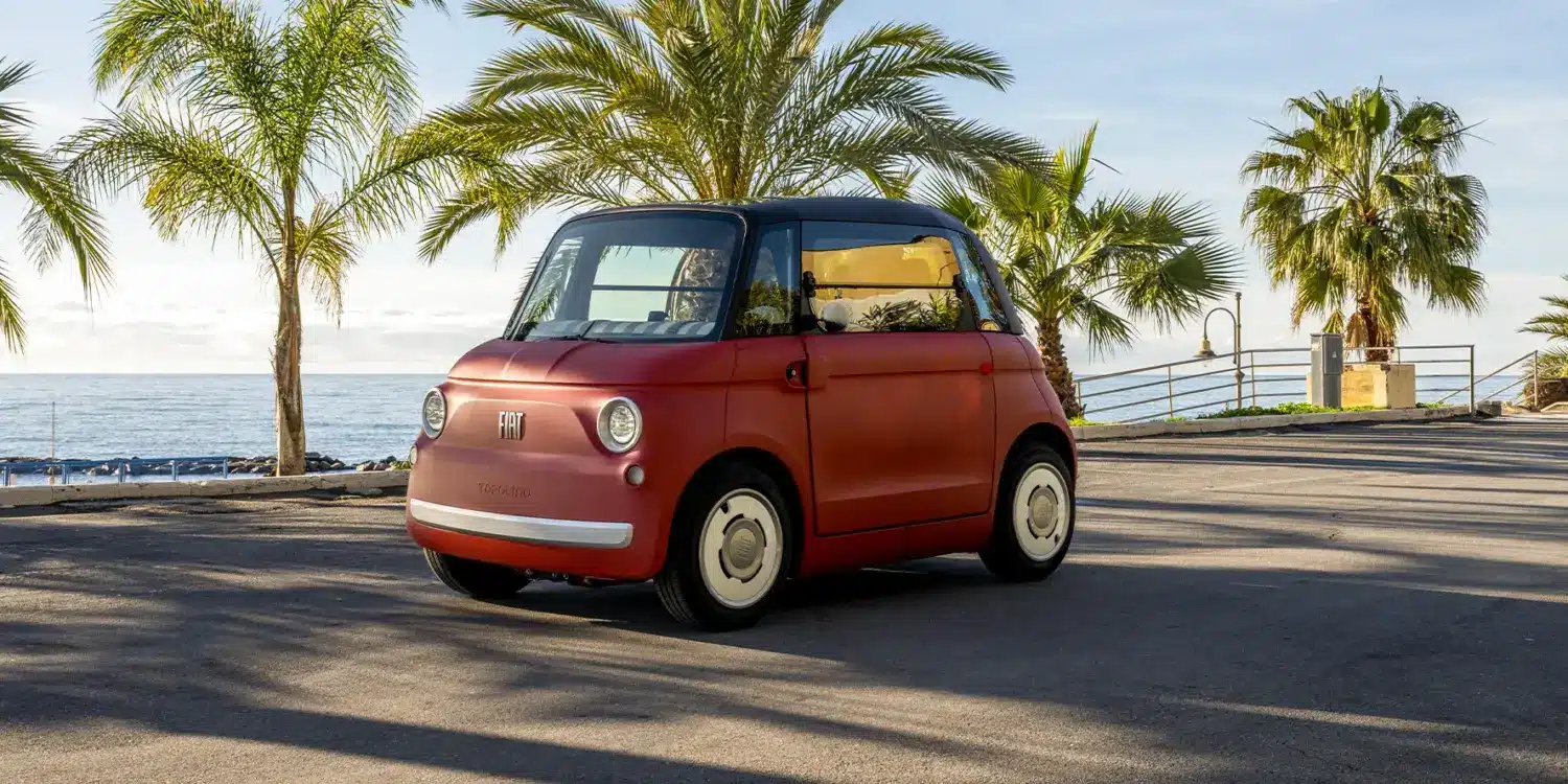 Fiat Topolino Microcar, vorne links, in Corallo vor Palmen und Meer geparkt.