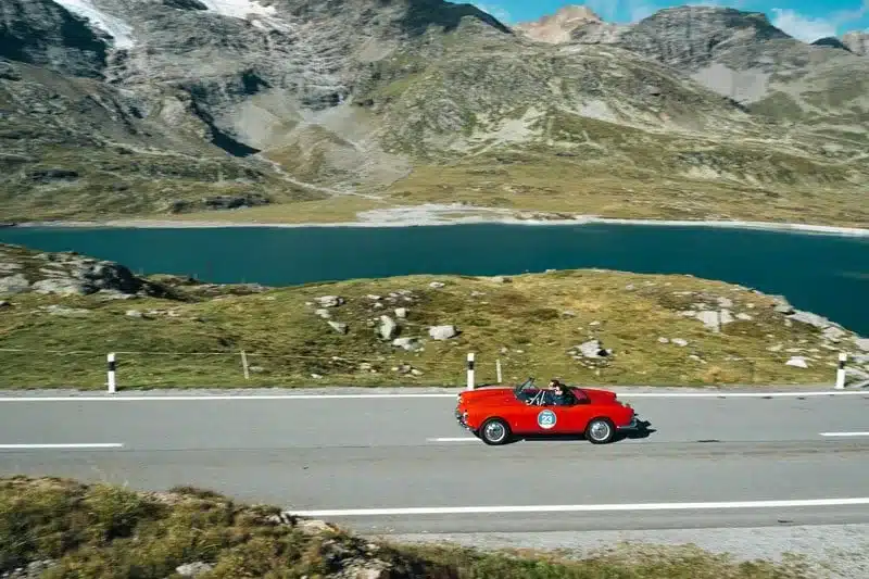 Klassisches rotes Cabrio fährt auf einer Passstraße mit Bergen und See im Hintergrund.