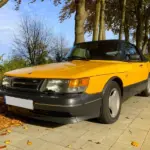 Gelber Youngtimer-Cabrio-Oldtimer steht auf herbstlicher Allee, gepflegter Zustand, klassische Linienführung.