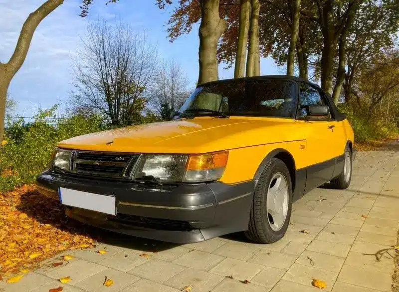 Gelber Youngtimer-Cabrio-Oldtimer steht auf herbstlicher Allee, gepflegter Zustand, klassische Linienführung.