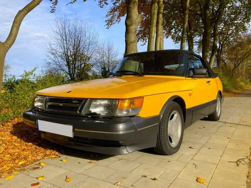 Gelber Youngtimer-Cabrio-Oldtimer steht auf herbstlicher Allee, gepflegter Zustand, klassische Linienführung.