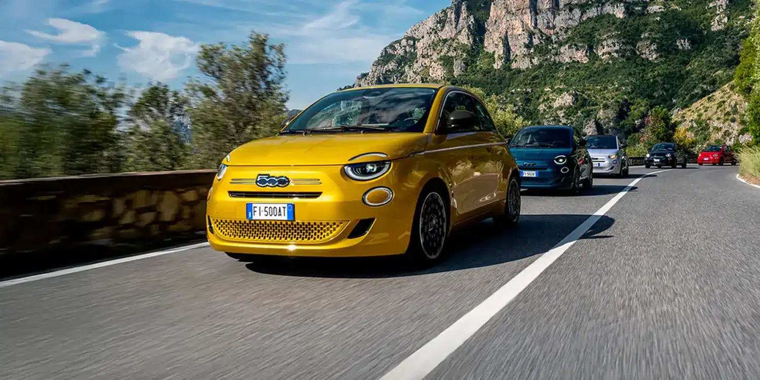 Mehrere Fiat 500 Hybrid fahren auf einer Bergstraße, vorn ein gelber Wagen, umgeben von üppiger Vegetation und Felslandschaft.