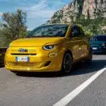 Mehrere Fiat 500 Hybrid fahren auf einer Bergstraße, vorn ein gelber Wagen, umgeben von üppiger Vegetation und Felslandschaft.