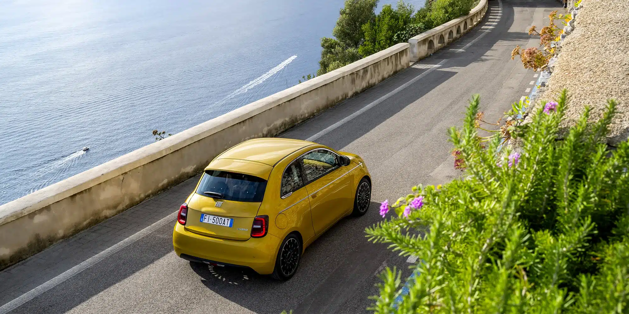 Fiat 500 Hybrid in Gelb fährt auf einer Straße an der Küste entlang, Meer im Hintergrund
