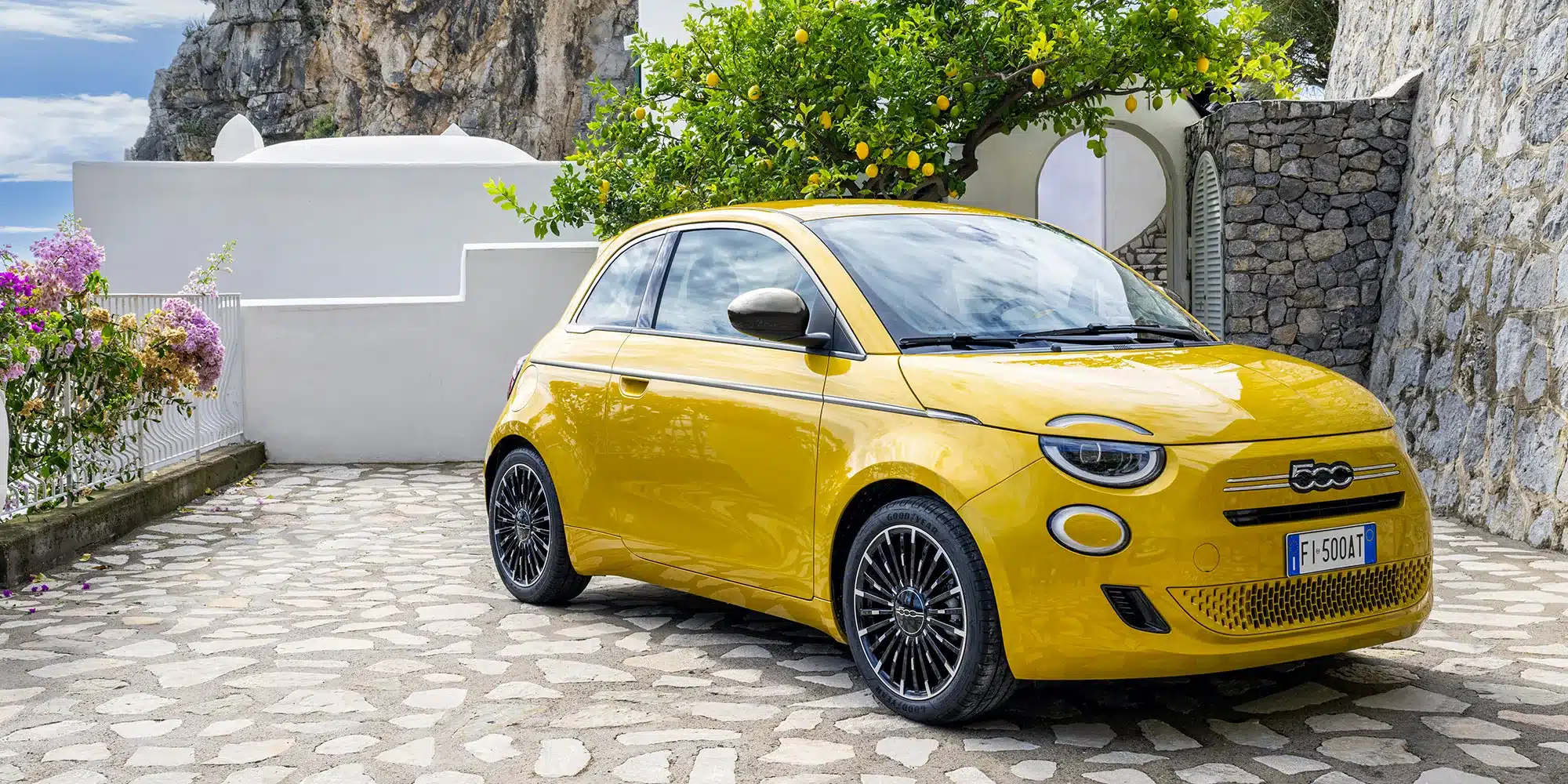 Gelber Fiat 500 Hybrid parkt auf gepflastertem Weg vor einer weißen Mauer und einem Zitronenbaum.