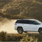 Weißer Jeep Grand Cherokee 4xe fährt auf einer staubigen Piste durch hügelige Landschaft.
