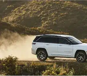 Weißer Jeep Grand Cherokee 4xe fährt auf einer staubigen Piste durch hügelige Landschaft.