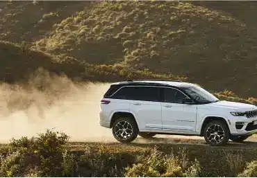 Weißer Jeep Grand Cherokee 4xe fährt auf einer staubigen Piste durch hügelige Landschaft.
