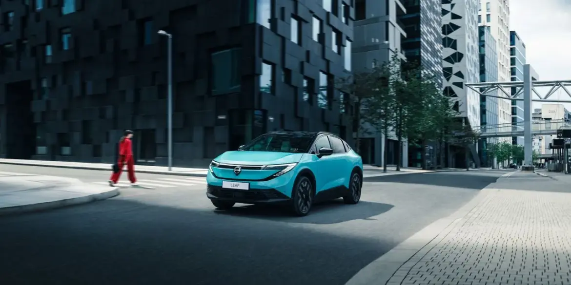 Nissan LEAF 2024 in türkis auf einer Straße vor modernen Hochhäusern.