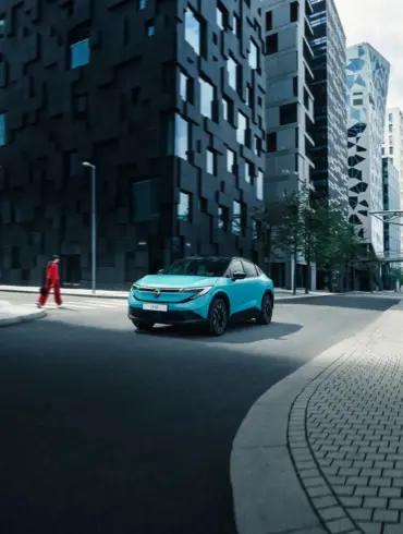 Nissan LEAF 2024 in türkis auf einer Straße vor modernen Hochhäusern.