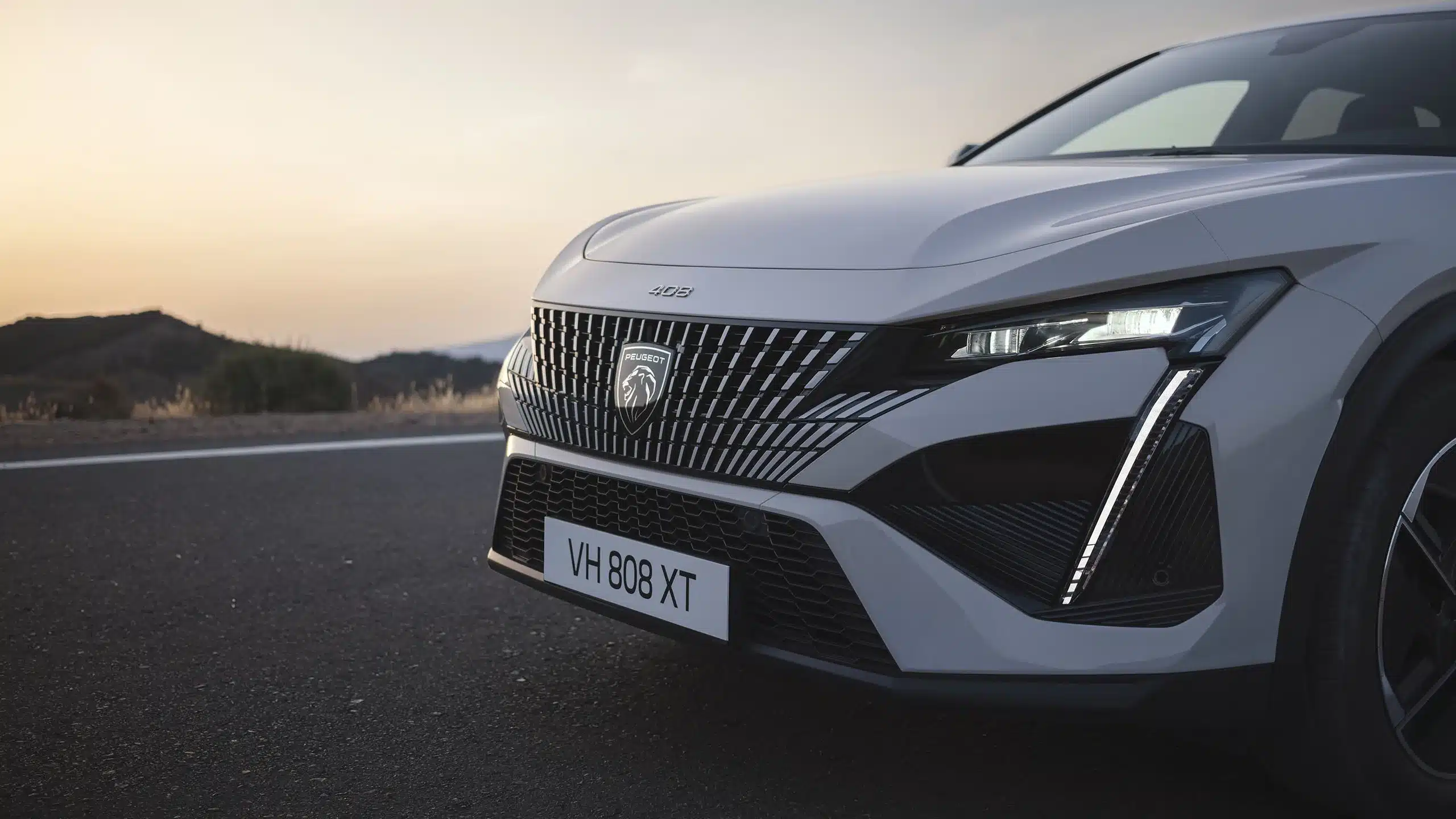 Die Frontpartie des Peugeot E-408 mit beleuchtetem Markenlogo, modernem Kühlergrill und LED-Tagfahrlicht vor einer Landschaft bei Sonnenuntergang.