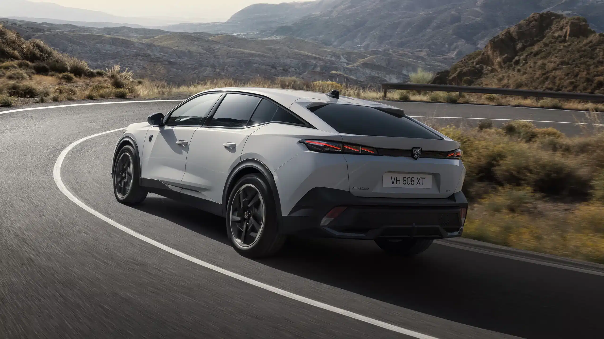 Peugeot E-408 fährt dynamisch auf einer Bergstraße, Seiten- und Heckansicht, weißes Auto vor Landschaft