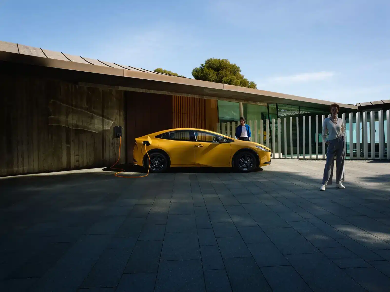 Gelber Toyota Prius Plug-in Hybrid lädt an einer Wallbox vor einem modernen Haus, zwei Personen stehen daneben.