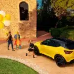 Gelber Nissan Juke mit schwarzem Dach vor Steinmauer und Personen mit gelben und weißen Ballons.