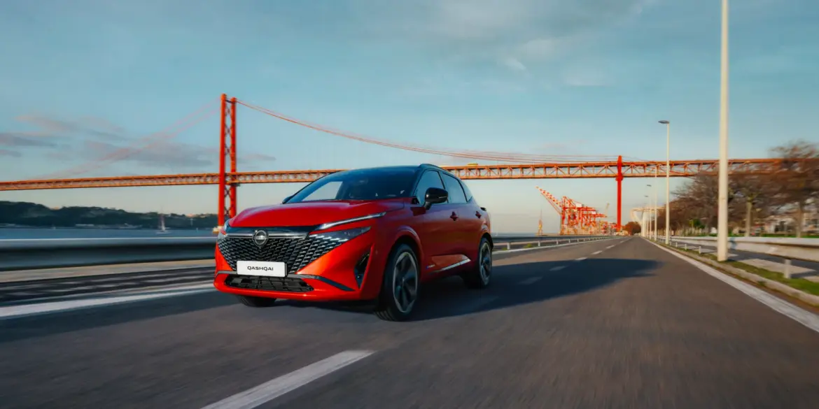Nissan Qashqai e-POWER in Rot fährt auf einer Straße mit Brücke im Hintergrund.