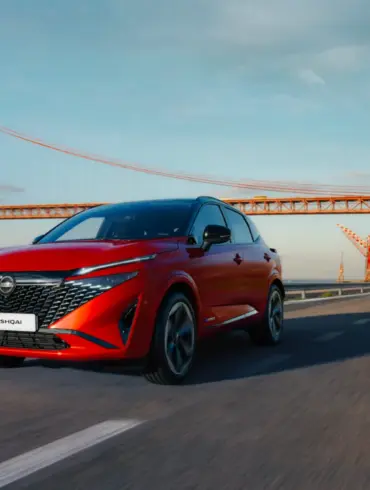 Nissan Qashqai e-POWER in Rot fährt auf einer Straße mit Brücke im Hintergrund.