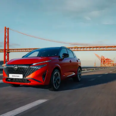 Nissan Qashqai e-POWER in Rot fährt auf einer Straße mit Brücke im Hintergrund.