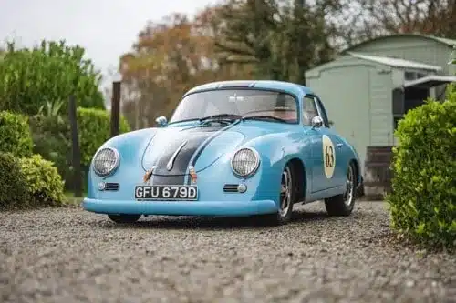 Blauer Porsche 356 Outlaw Tribute mit Streifen und Startnummernkreisen, steht auf einem Kiesweg.