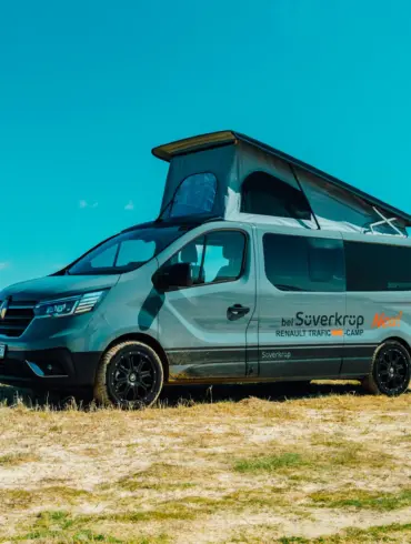 Renault Trafic BS-Camp Campervan mit geöffnetem Aufstelldach steht auf einer Wiese unter blauem Himmel