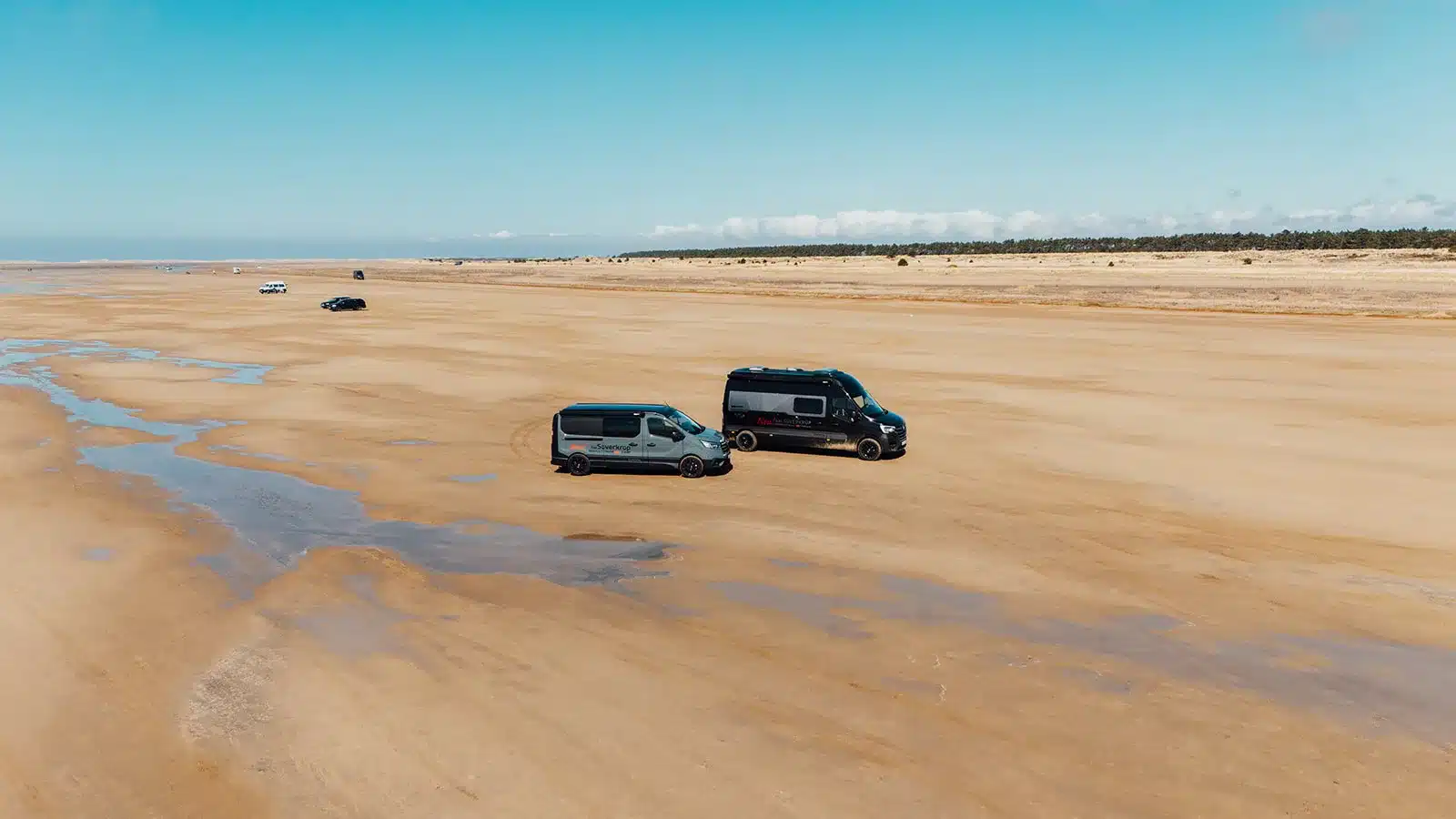 Zwei Camper-Vans, darunter der Renault Trafic BS-Camp, parken auf einer offenen Sandfläche mit weitem Horizont und blauem Himmel.