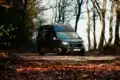 Renault Trafic BS-CAMP Campervan fährt auf einem Laub bedeckten Weg durch herbstlichen Wald.