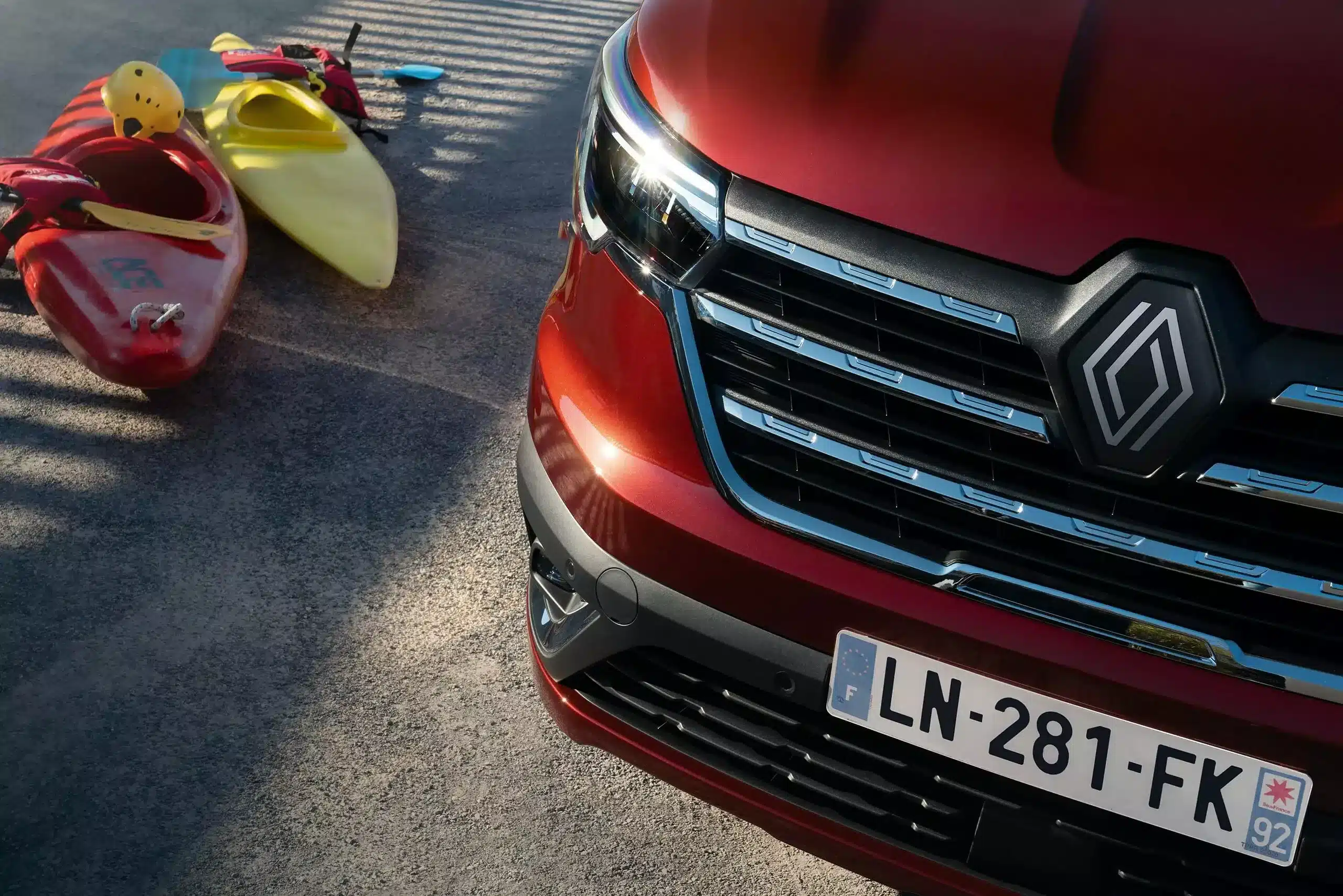 Front des Renault Trafic in Rot mit zwei Kajaks im Hintergrund auf einem Parkplatz.