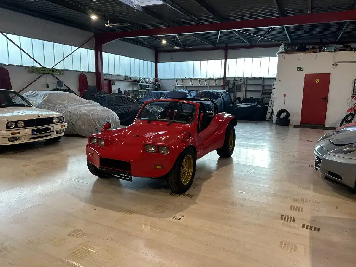 Roter Volkswagen Deserter GT Buggy von 1970 mit goldenen Felgen, Aufnahme in einer hellen Garage zwischen klassischen Sportwagen.