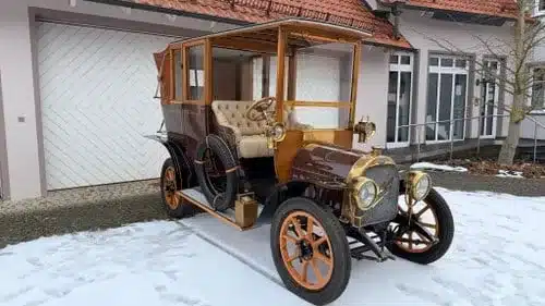 Ein restaurierter klassischer Oldtimer mit Holzkarosserie steht auf schneebedecktem Boden vor einem Wohnhaus
