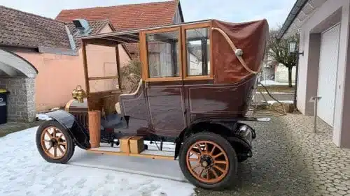 Eleganter Oldtimer mit Holzspeichenrädern, abgerundeter Karosserie und Lederdach vor einem Haus geparkt.