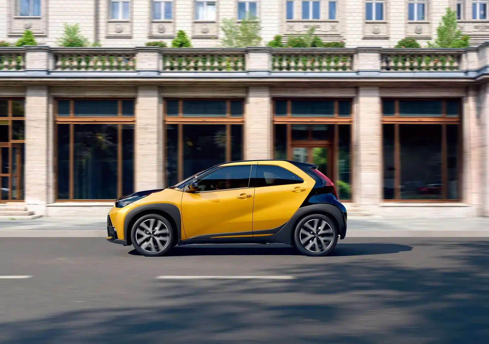 Toyota Aygo X Hybrid fährt auf städtischer Straße vor klassischer Gebäudefassade.