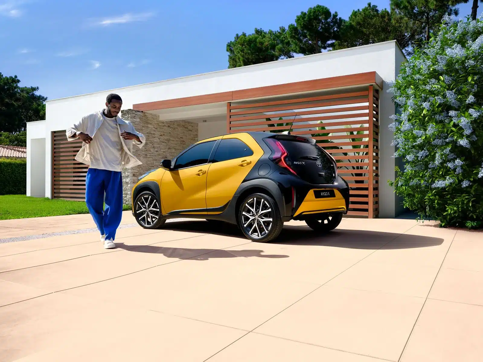 Toyota Aygo X Hybrid in Gelb-Schwarz vor einem modernen Haus mit Garten, daneben eine Person.